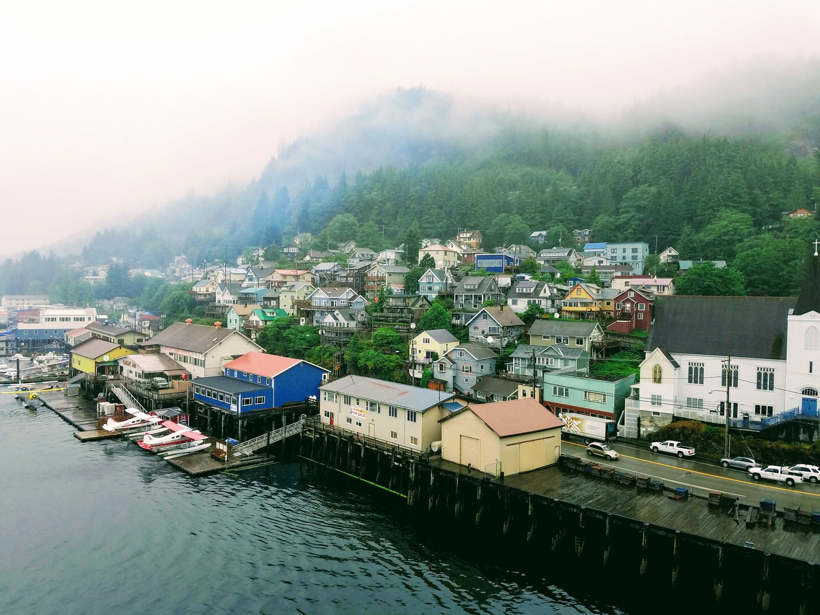 Ketchikan waterfront seen from a cruise ship at dawn, painted wooden buildings on pilings below a fog-covered green hillside