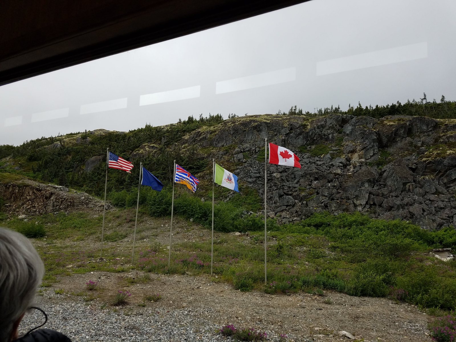 Four flags flying at the White Pass summit on bare rocky ground, marking the United States and Canada border