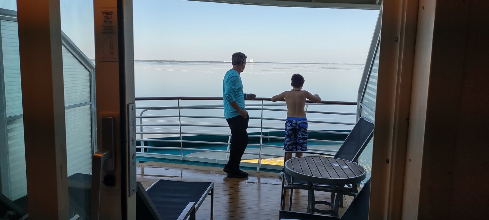 Two people at a cruise ship balcony rail, seen from inside the cabin, looking out at a calm sea