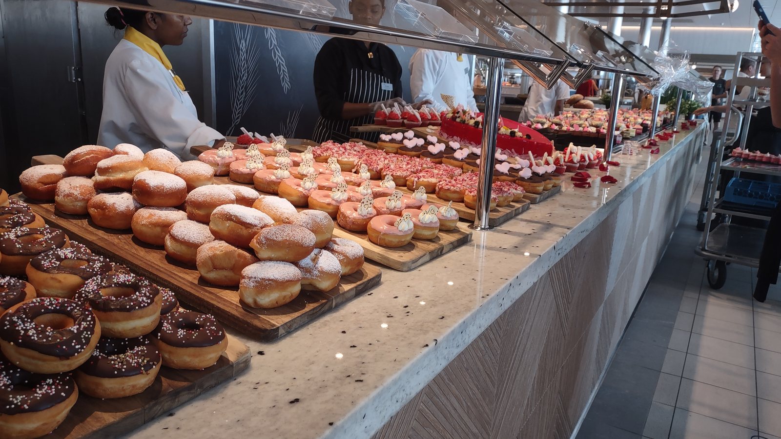 A cruise ship buffet dessert counter, trays of doughnuts and pastries with chefs behind the glass