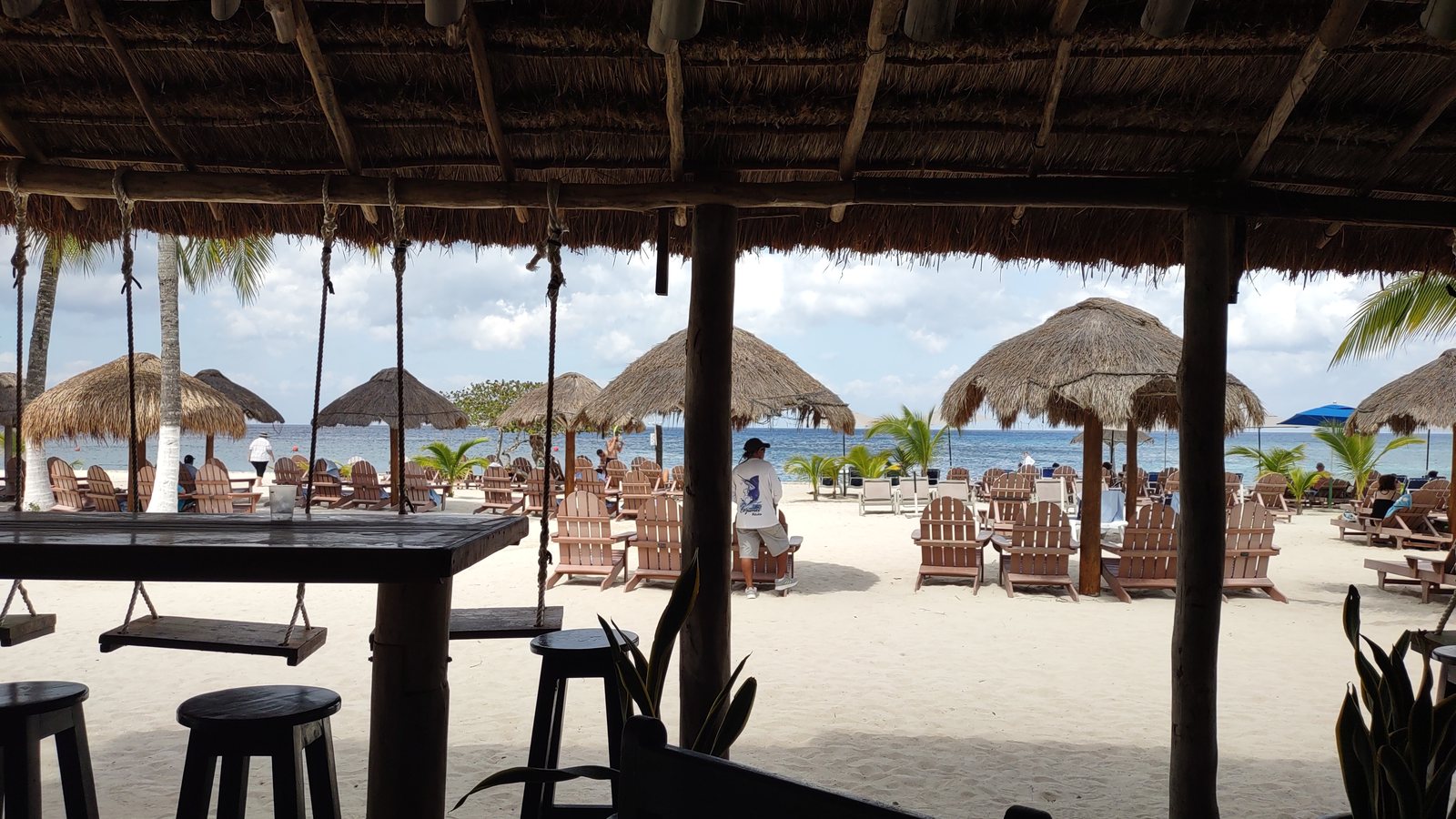 A palapa beach bar in Cozumel with swing seats hanging at the counter, looking out past thatched umbrellas to the sea