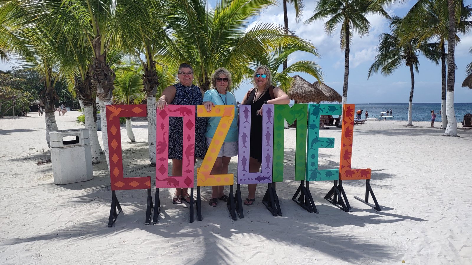 Three women standing behind the big painted COZUMEL letters on the beach, palm trees and turquoise sea behind them