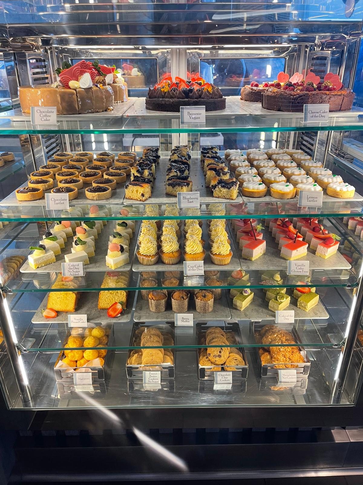 A refrigerated dessert display case on a cruise ship filled with rows of small cakes and pastries