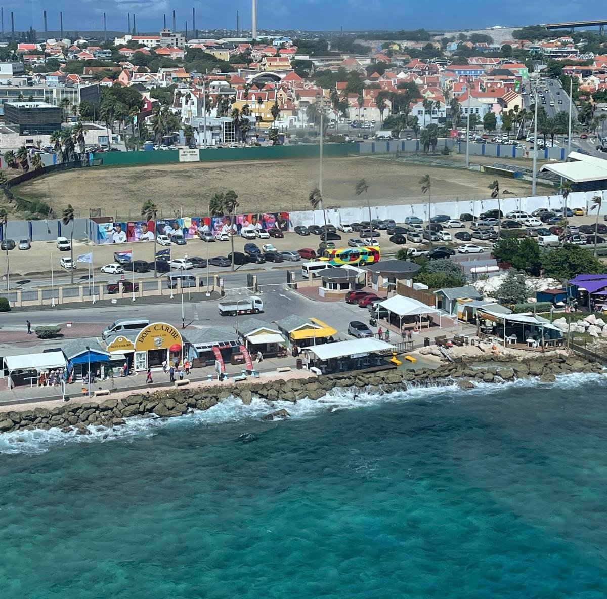Looking down from a cruise ship at the Oranjestad waterfront in Aruba, a row of small shops and awnings along a seawall with people walking and the sea breaking on the rocks below