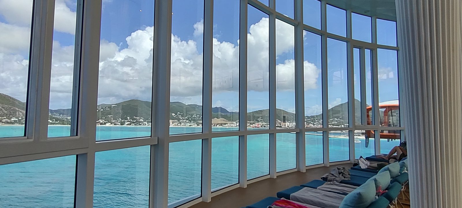 A curved wall of windows in a ship lounge looking out at a Caribbean island, cushioned seating along the glass