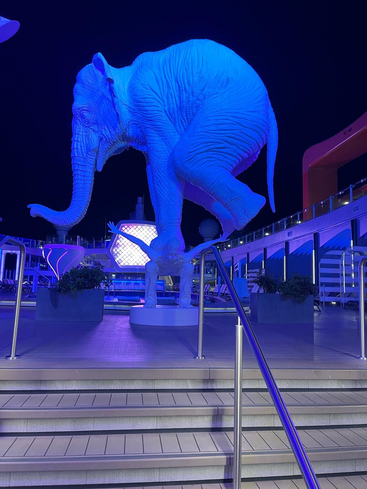 A large illuminated elephant sculpture glowing blue on a ship deck at night, surrounded by purple lighting
