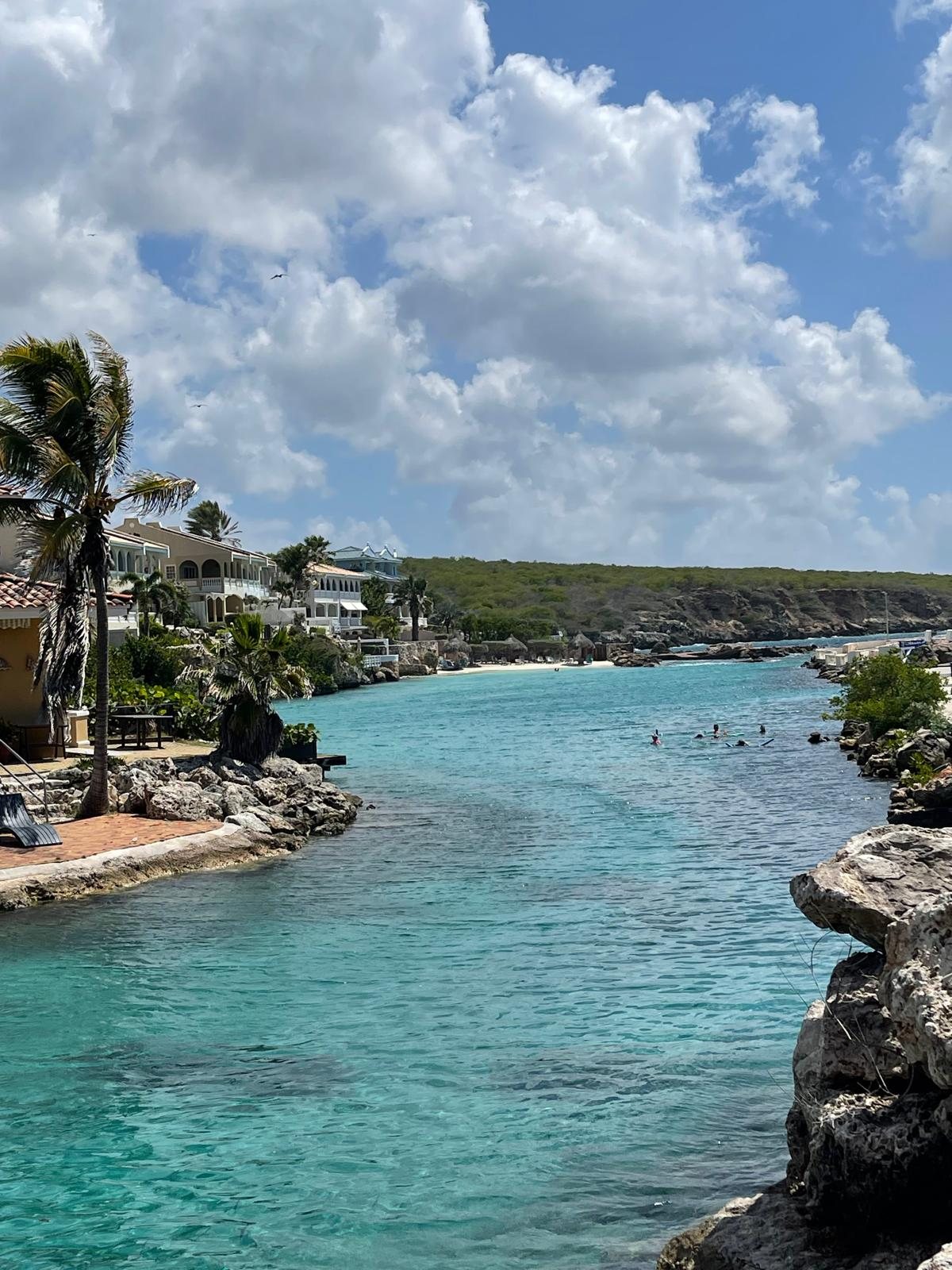 A narrow turquoise inlet in Curacao framed by low limestone rock, with villas and palms along the left shore and swimmers in the water